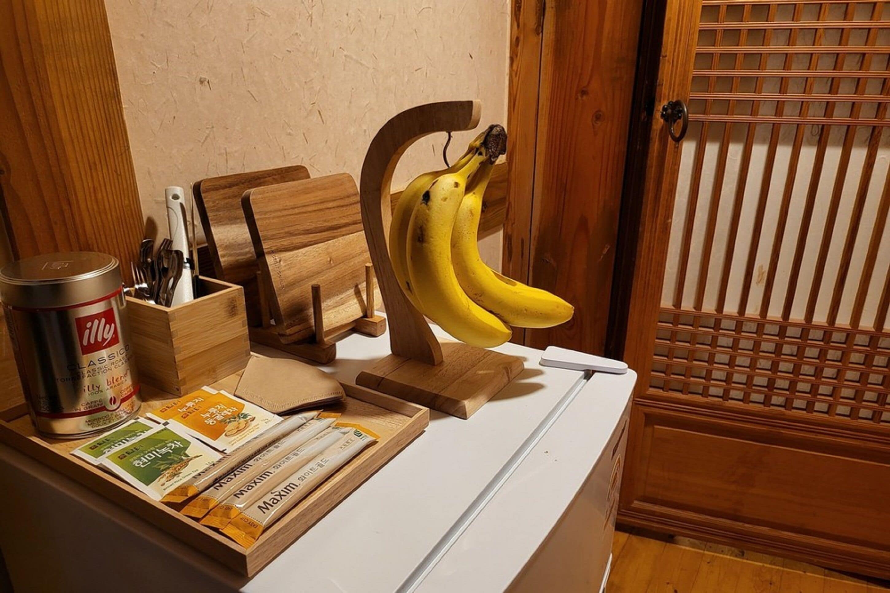 Een keukentafel met een houten fruitmand, gevuld met bananen, een blik Illy koffie, een messenblok en een paar receptenkaarten.