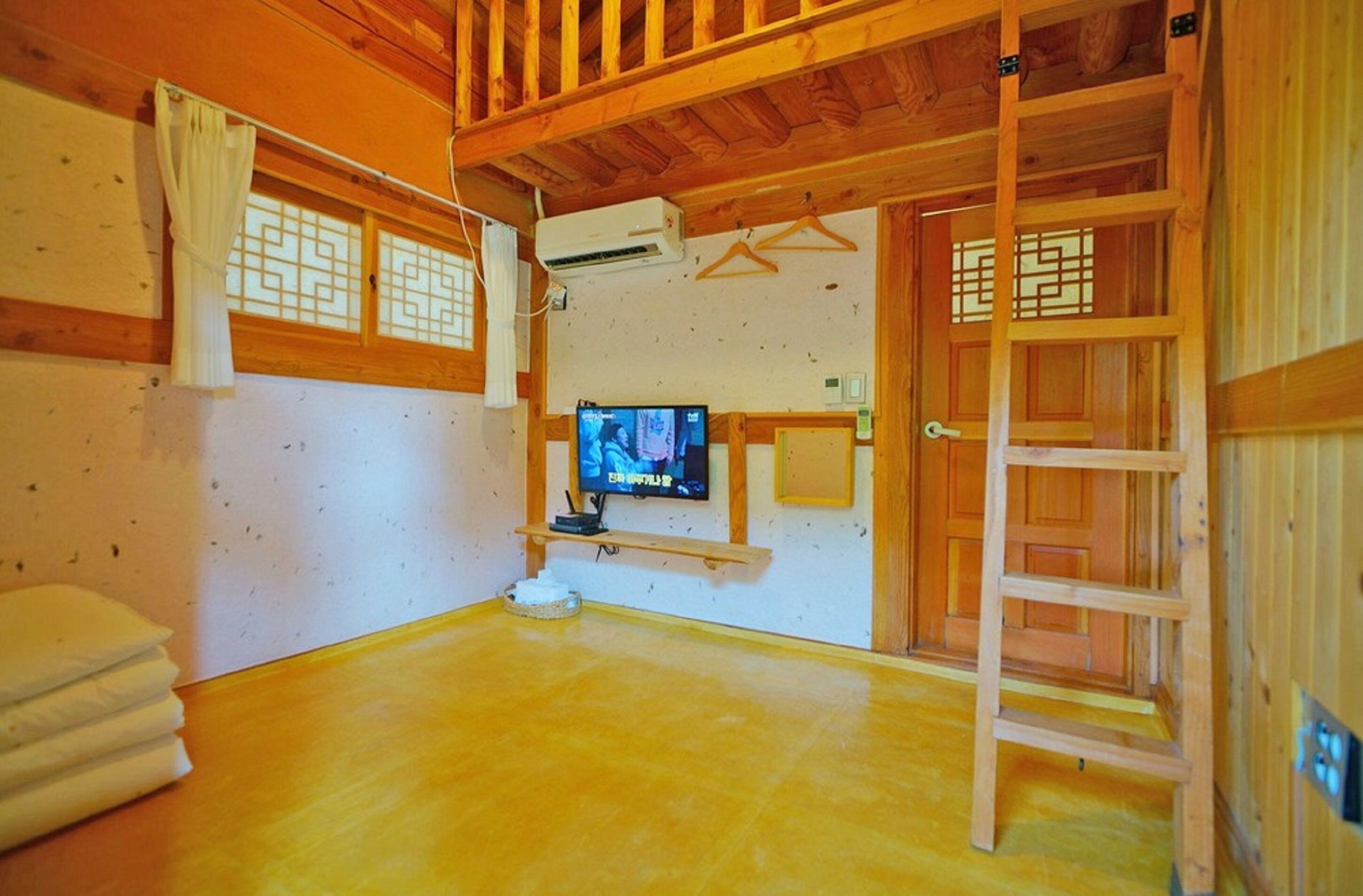 Une pièce avec un plancher en bois, un téléviseur posé sur un meuble, une échelle et un rideau blanc.