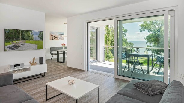 Ein modernes Wohnzimmer mit einer Couch, einem Couchtisch und einem an der Wand montierten Flachbildfernseher. Eine Glastür führt zu einem Außenbalkon mit Tisch und Stühlen.