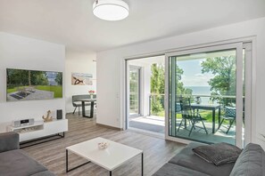Moderni olohuone, jossa on sohva, sohvapöytä ja seinälle asennettu taulutelevisio. Huoneesta on liukuovet ulos avautuvalle parvekkeelle, jossa on pöytä ja tuolit.