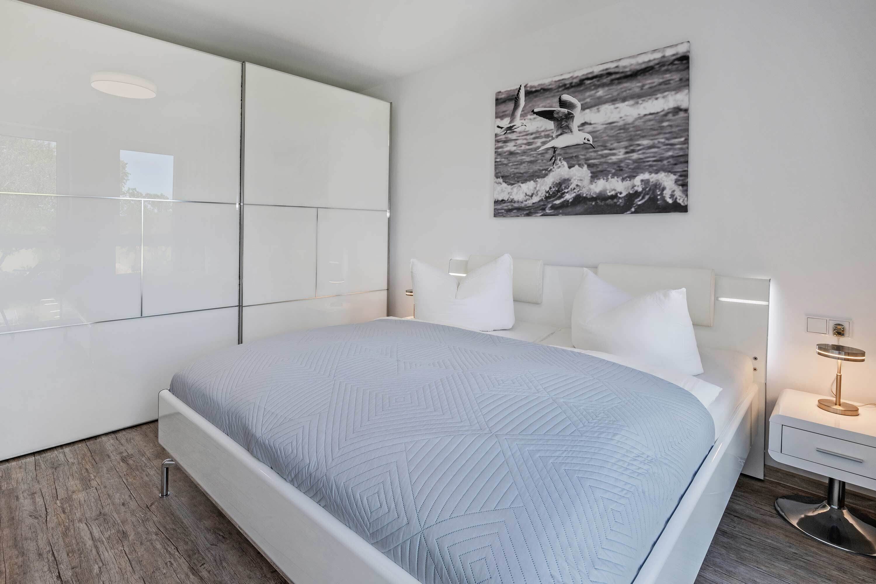 A modern bedroom with a large bed, a bedside table with a lamp, a wall-mounted artwork, and a large wardrobe.