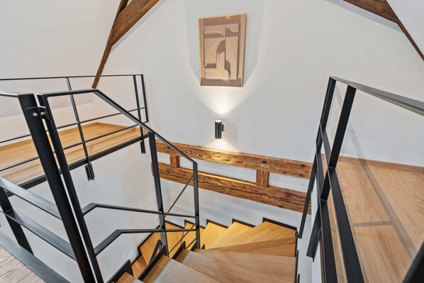 Una escalera moderna con escalones de madera y barandillas de metal, que conduce a un rellano con una lámpara fijada a la pared.