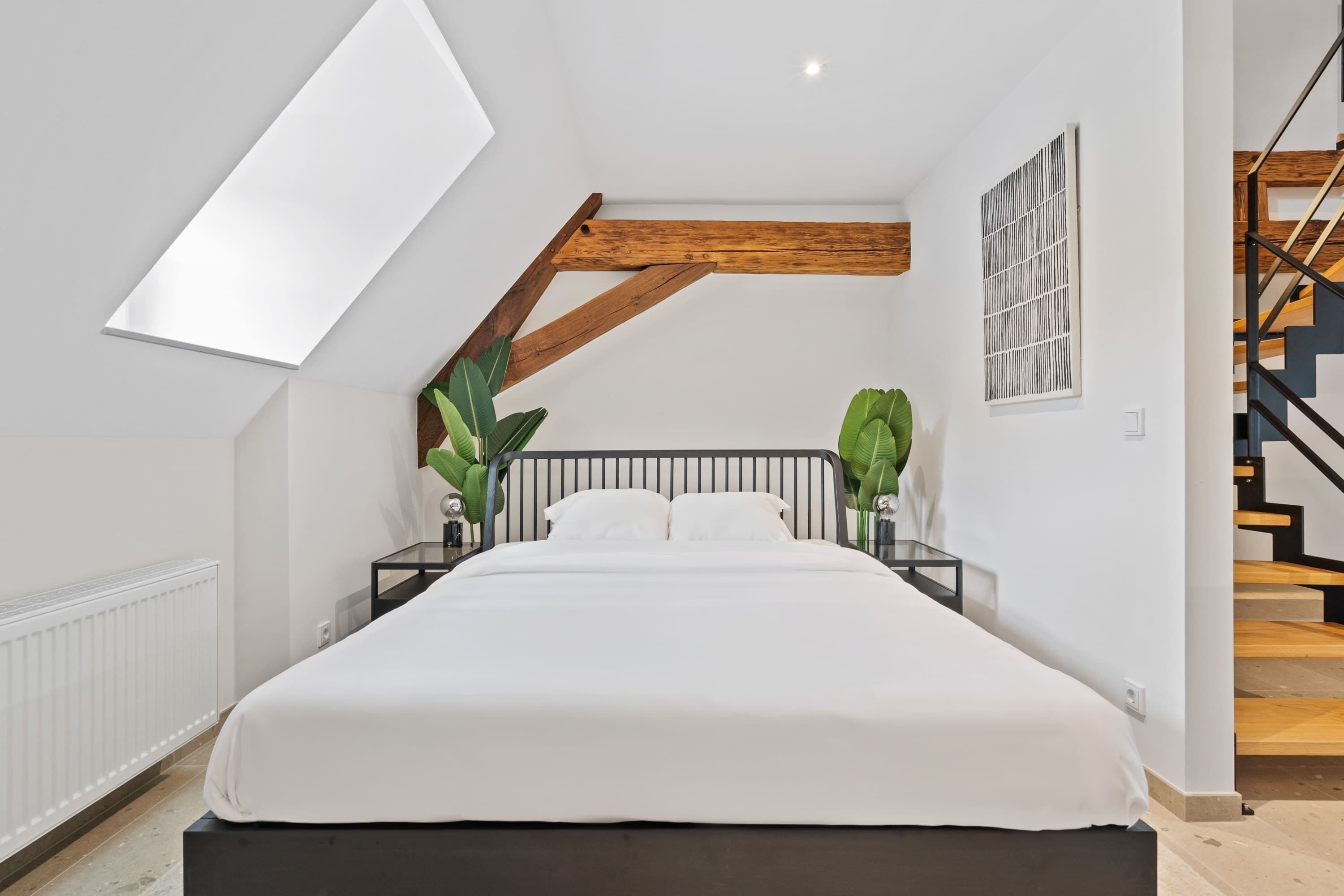 Un dormitorio con una cama grande, techo inclinado, viga de madera, radiador, mesita de noche negra, pared blanca y una escalera.