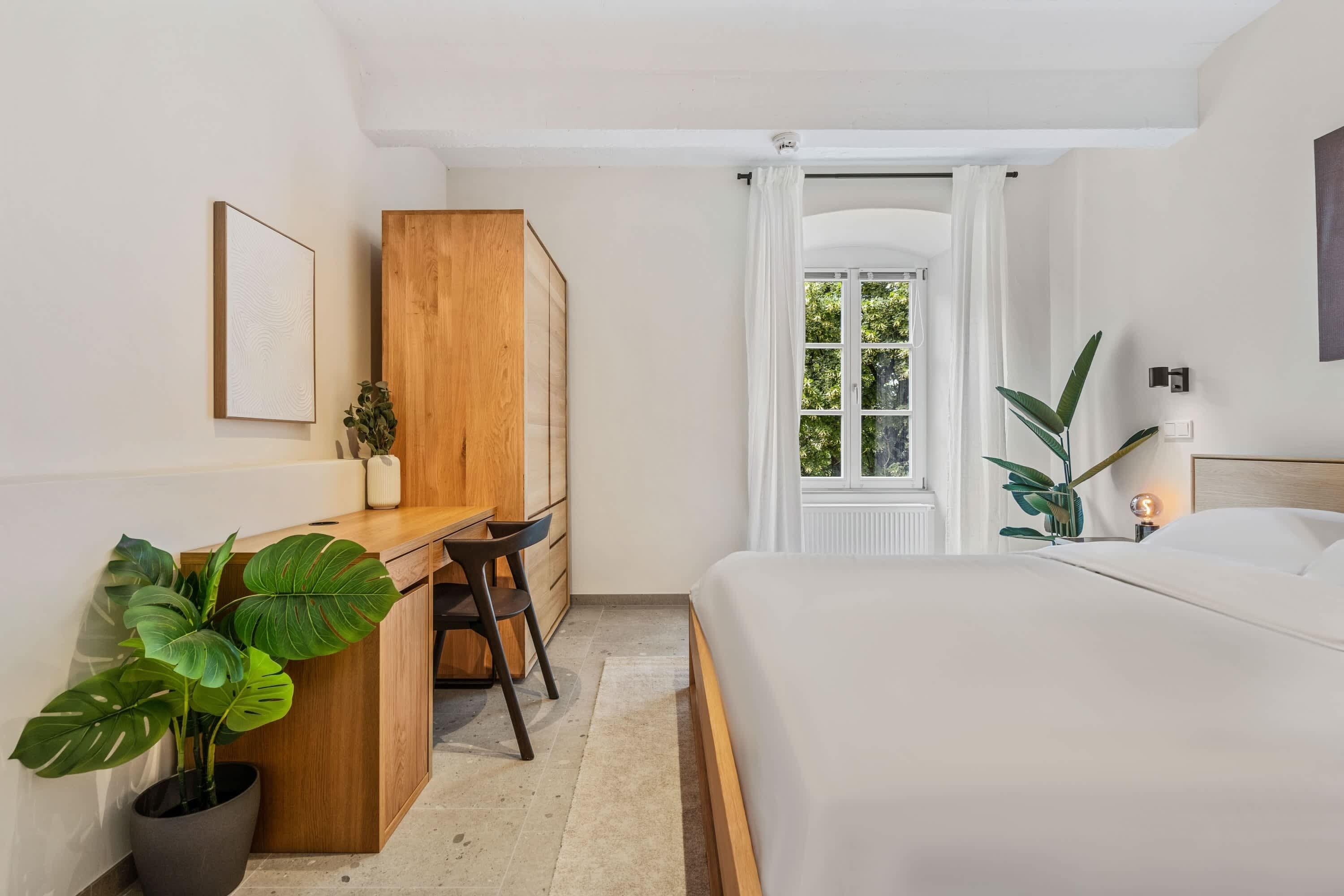 A bedroom with a bed, a desk, a chair, a potted plant, and a window with curtains.