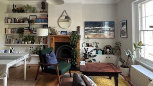 Een gezellige woonkamer met een open haard, boekenplanken, een groene fauteuil, een houten salontafel en een groot raam.