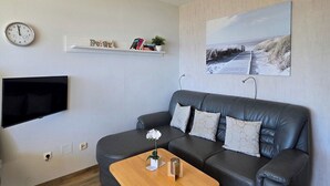 Een moderne woonkamer met een grijze leren bank, een houten salontafel, een wandgemonteerde flatscreen televisie, een plafondlamp, een wandklok, een plank met boeken en een klein plantje, en een schilderij met een strandthema.