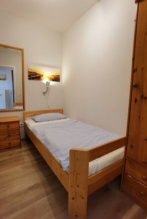Ein kleines, ordentlich eingerichtetes Schlafzimmer mit einem Einzelbett, einer Holzkommode, einem Nachttisch und einer Wandleuchte.