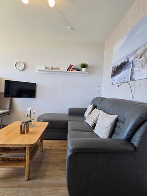 Ein modernes Wohnzimmer mit grauem Ledersofa, einem Holztisch, einem Flachbildfernseher, einem an der Wand befestigten Regal mit Büchern und einer Pflanze sowie einem großen Landschaftsbild.