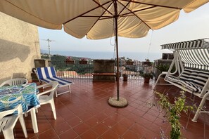 Eine Terrasse mit Tisch und Stühlen, ein gestreifter Liegestuhl, ein runder Tisch und ein großer Sonnenschirm.