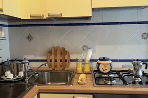 Een keuken met een gootsteen, fornuis en diverse keukenbenodigdheden, zoals een koffiezetapparaat en een houten snijplank.