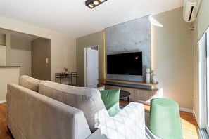 A modern living room with a large flat-screen TV mounted on a textured wall, a grey sofa with green cushions, a small round table, and a large window providing natural light. - Curated 1BR Design Apartment in Palermo Soho · Hosted by Welcome2BA (Palermo)