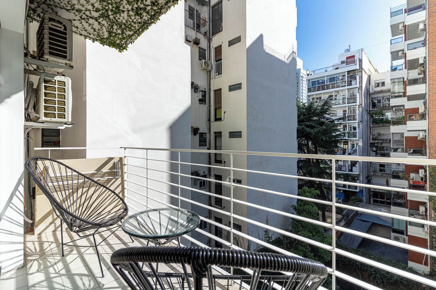 Un balcon avec deux chaises en fil de fer noir et une table en verre, donnant sur un quartier résidentiel composé de plusieurs immeubles d’appartements.