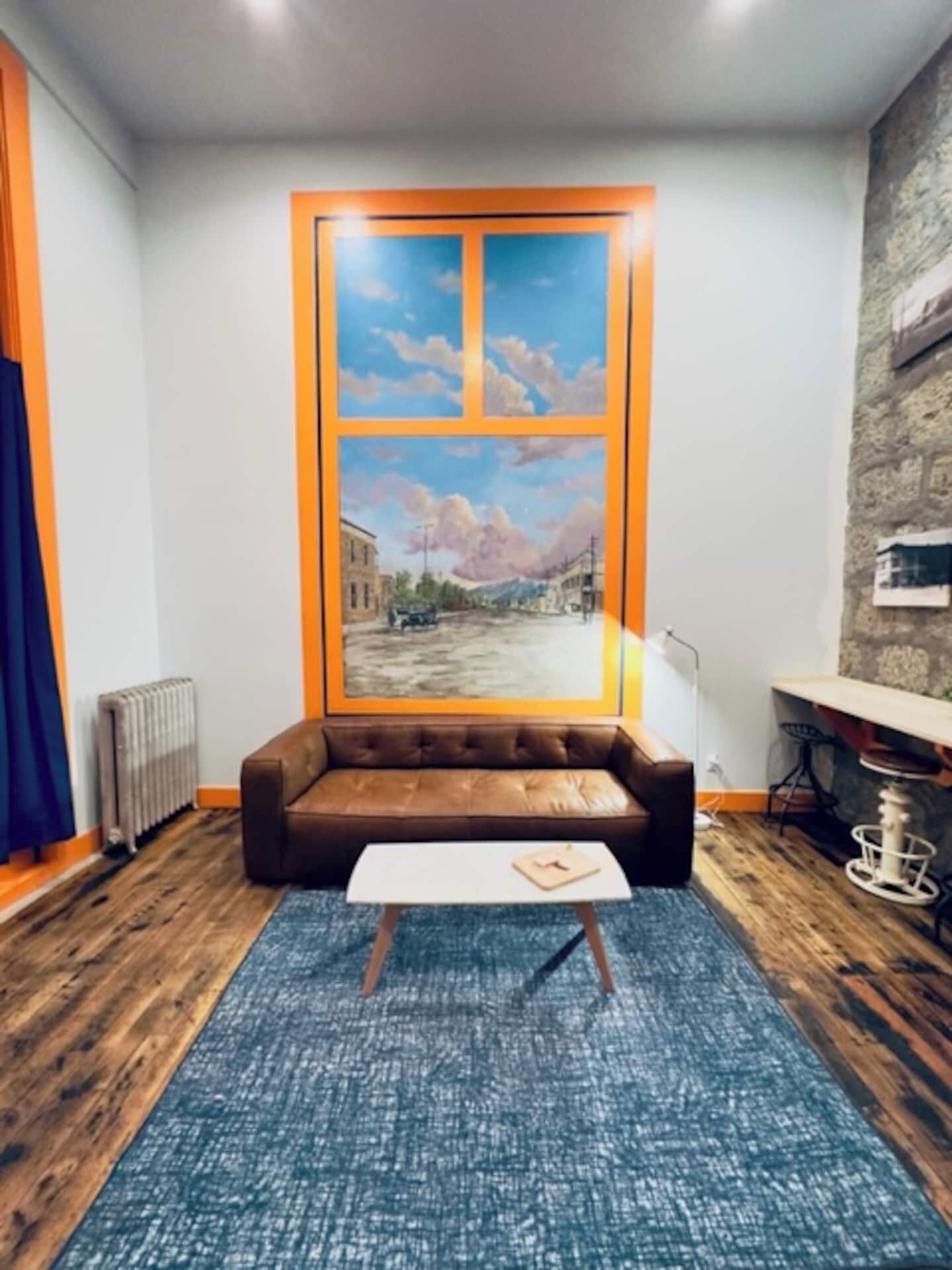 Una habitació amb un sofà de cuir marró, una taula de cafè blanca, una catifa blava i un gran finestral amb vista a la ciutat.