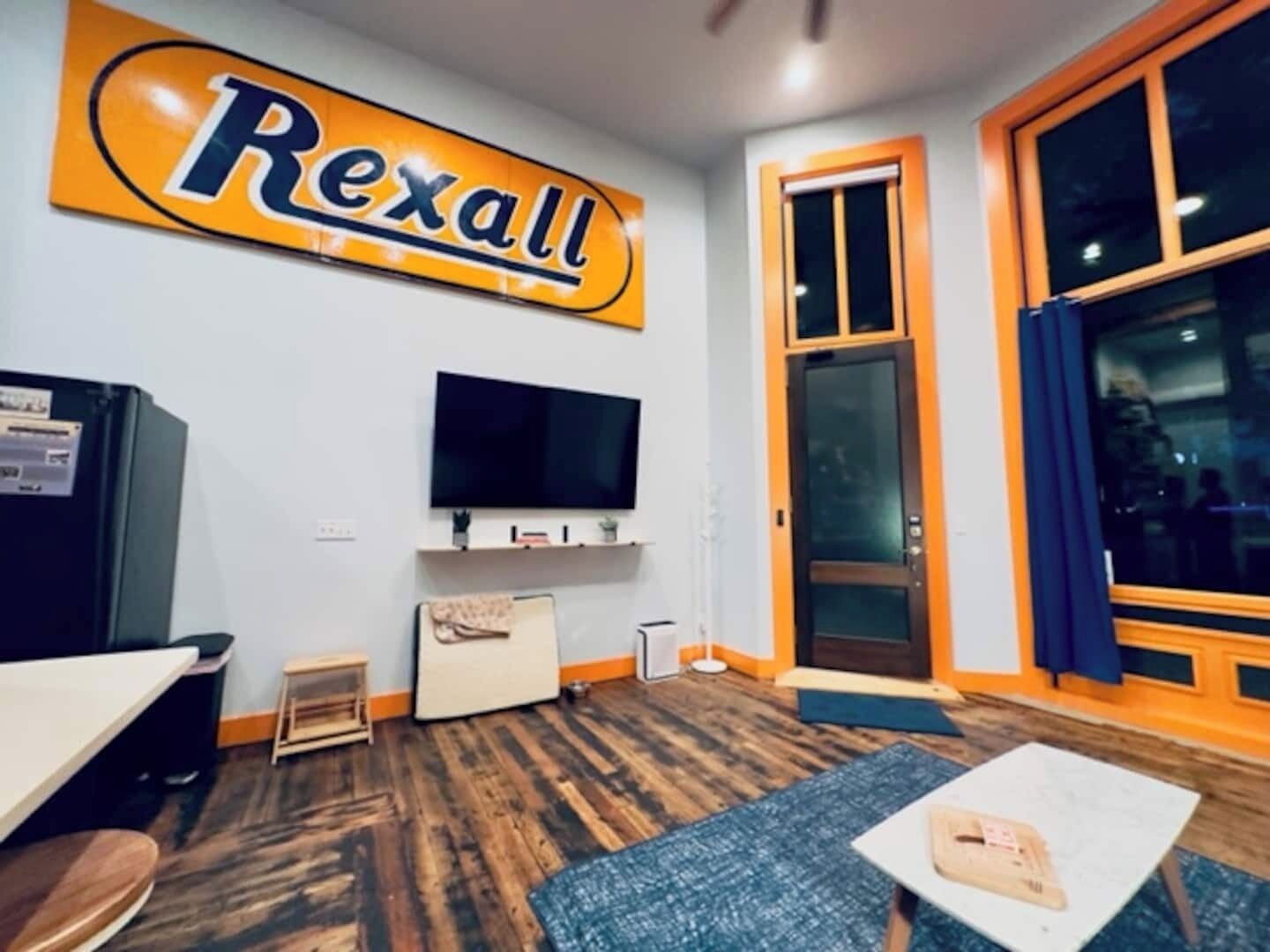A room with a large Rexall sign, a flat-screen TV, a small table, and a window with a view.