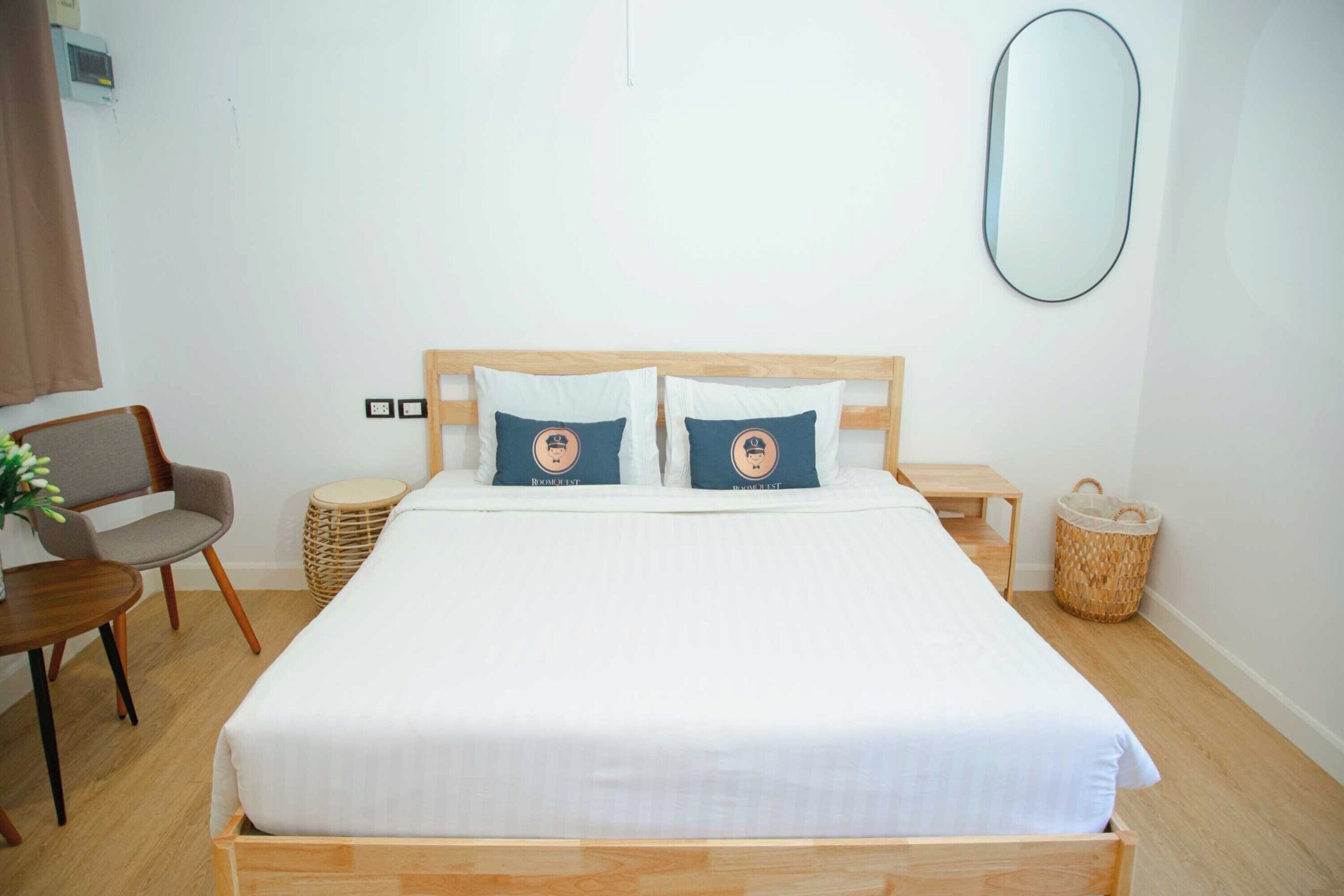A neatly arranged bedroom with a wooden bed, two pillows, a bedside table, a chair, a small round table, and a woven basket.