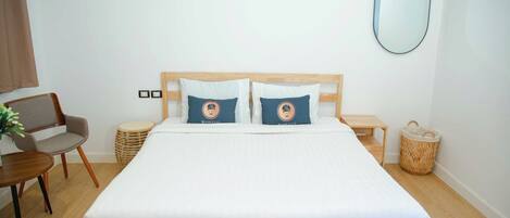 A neatly arranged bedroom with a wooden bed, two pillows, a bedside table, a chair, a small round table, and a woven basket.