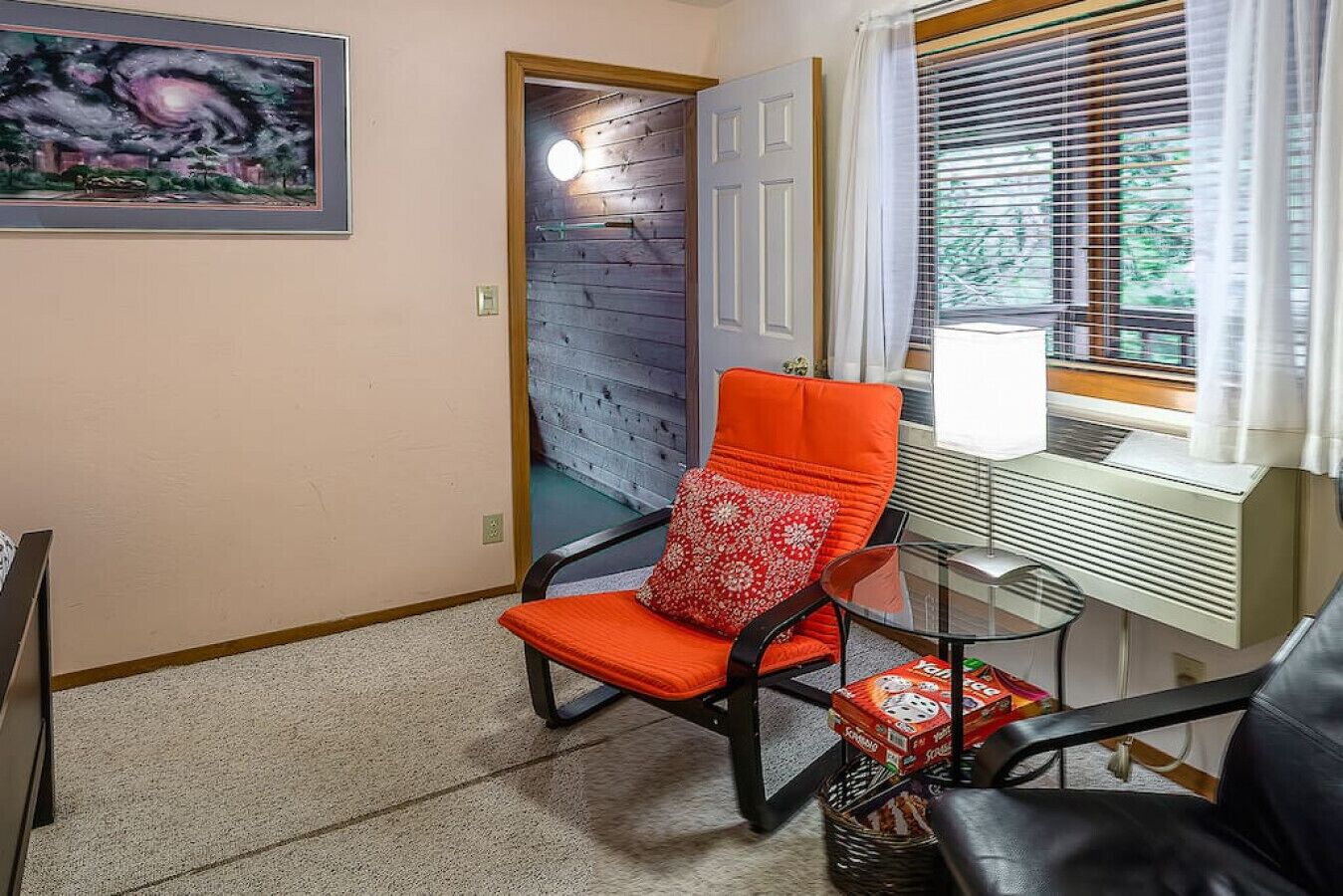 A room with a rocking chair, a desk with a lamp, a window with blinds, and a painting on the wall.