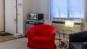 Un espace de vie compact comprenant un fauteuil rouge, une petite table, un téléviseur et une fenêtre avec des stores.