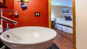 A bathroom with a large white sink, a red wall, and a bed with a floral bedspread in the background. - Lodge Room 5 with Jacuzzi Tub at Green Springs Inn (Ashland)