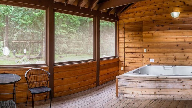 Uma cabana de madeira com uma janela grande, uma banheira de hidromassagem e uma mesa com cadeiras.