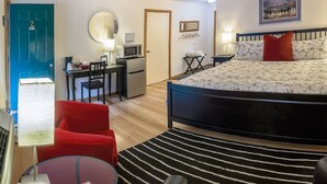 Quarto de hotel compacto com cama, escrivaninha com computador, cadeira, pequeno espaço de cozinha e televisão.