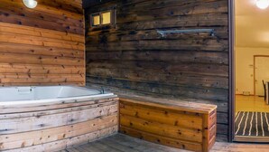 Interior de uma cabana de madeira com uma banheira, paredes de madeira e uma porta que dá para outro quarto.
