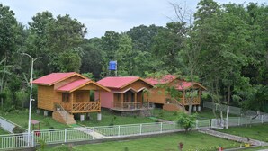 A row of wooden cabins with red roofs, surrounded by lush greenery and a paved walkway. - De Aloha Resort (Tezpur)