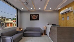 A modern reception area with a grey sofa, a wooden reception desk, and a large window offering a view of the street. - Hotel Emporio (Ghaziabad)