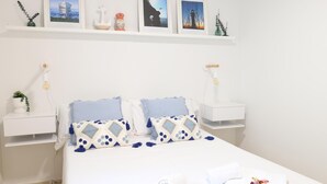 A neatly arranged bedroom with a bed, two bedside tables, a wall-mounted shelf with framed pictures, and a small potted plant.