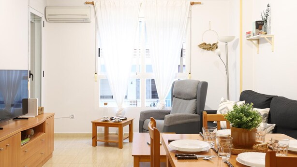 A modern living room with a sofa, a dining table set for two, a TV on a wooden cabinet, and a large window with sheer curtains.