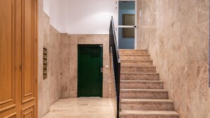 A marble staircase leading to a green door, with a wooden door on the left and a wall-mounted elevator control panel.