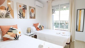 A hotel room with two beds, a small table with a lamp, a window with curtains, and artwork on the wall.