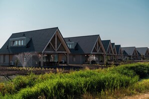 Une rangée de maisons modernes à plusieurs étages, dotées de toits à pignon et de grandes fenêtres, s’étend dans un espace aménagé, caractérisé par une végétation luxuriante.