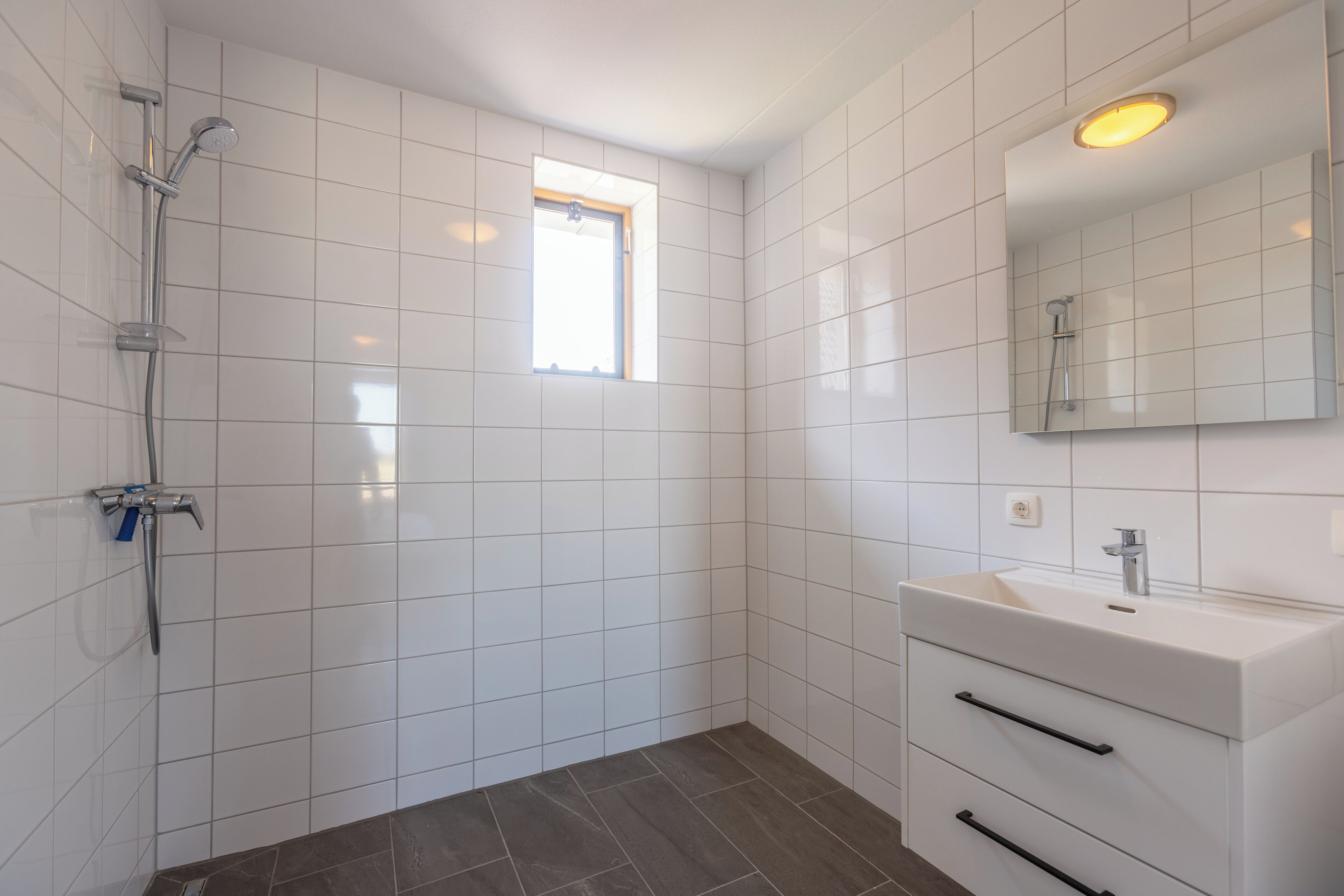 Uma casa de banho com paredes revestidas a mosaico branco, chuveiro, lavatório com espelho e janela.