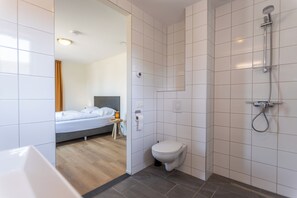 Une salle de bain moderne avec douche, toilettes et une chambre visible, équipée d’un lit et d’une table de chevet.