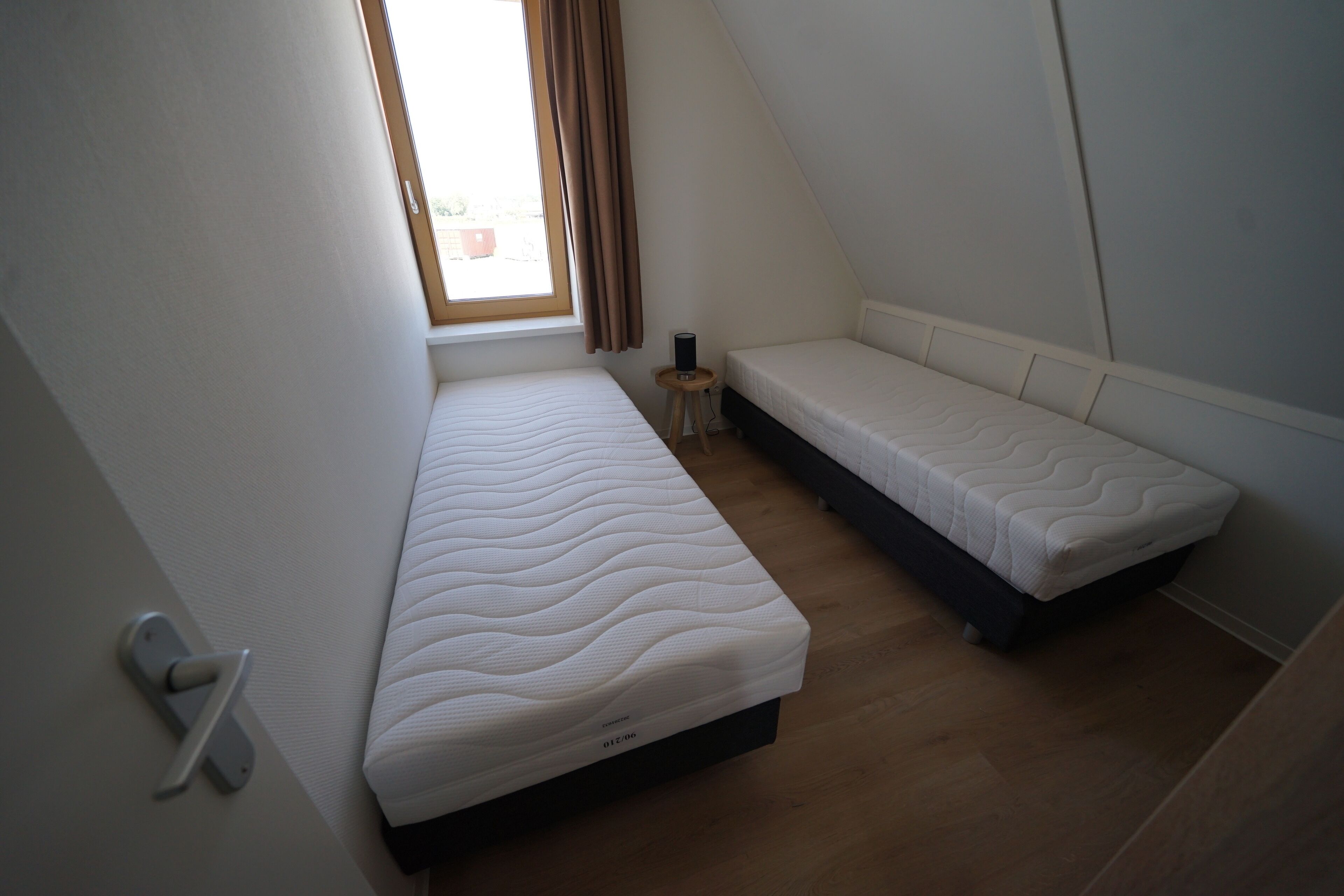 Habitació petita i moderna amb dos llits individuals, una taula petita i una finestra amb cortines.