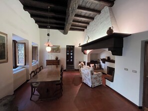 A spacious dining area with a long wooden table and chairs, a hanging light fixture, and a fireplace with a shelf above.