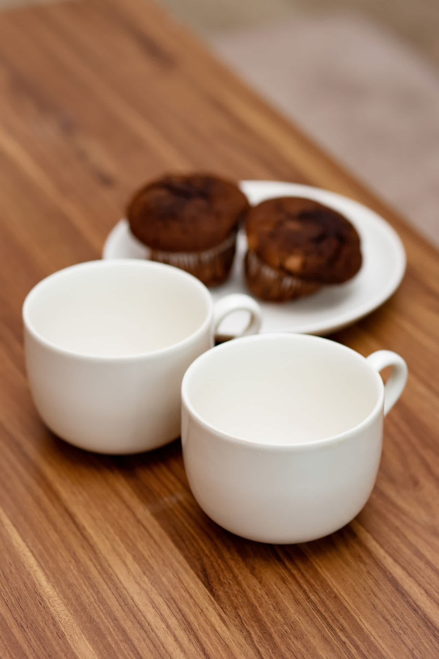Two white ceramic cups on a wooden table with a plate of muffins in the background.