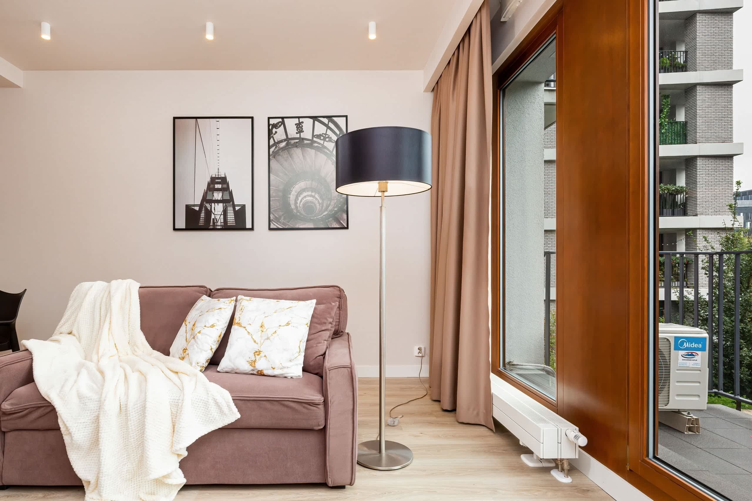 Een moderne woonkamer met een roze bank, een witte deken, twee decoratieve kussens, een vloerlamp en een groot raam met gordijnen.
