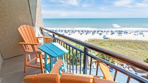 Ein Balkon mit Holzstühlen und einem Tisch, der auf einen Strand mit Sonnenschirmen und Menschen blickt.