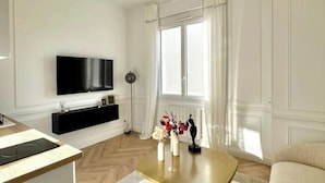 Een moderne woonkamer met een flatscreen televisie, een zwarte mediakast, een gouden salontafel met decoratieve objecten, een beige bank en een groot raam met witte gordijnen.