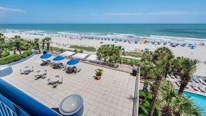A rooftop terrace with sun loungers and umbrellas, overlooking a beach with umbrellas and people. - Nice Oceanfront 1BR with Heated Pools, Coral Beach (Myrtle Beach)
