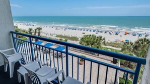 A balcony with white plastic chairs and a view of a beach with umbrellas and palm trees. - Nice Oceanfront 1BR with Heated Pools, Coral Beach (Myrtle Beach)