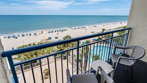 A balcony with a view of a beach, a pool, and a few chairs.