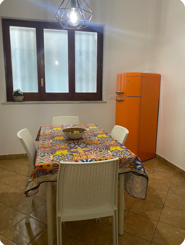Un coin repas avec une table recouverte d’une nappe colorée, entourée de chaises blanches, une fenêtre à vitres dépolies et un réfrigérateur orange.