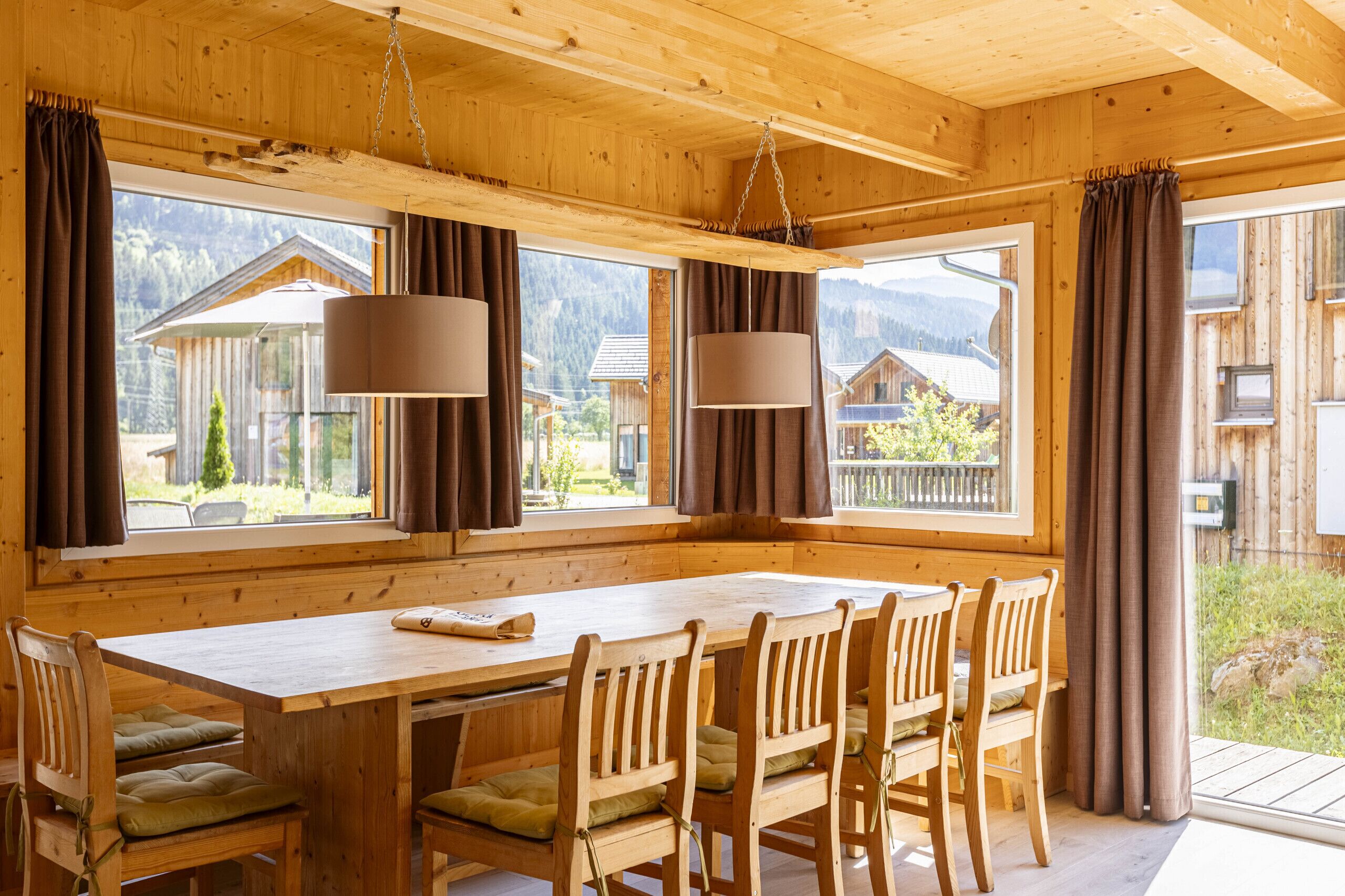 A wooden cabin with a large dining table and chairs, large windows with curtains, and a view of surrounding buildings and mountains.