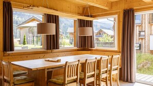 A wooden cabin with a large dining table and chairs, large windows with curtains, and a view of surrounding buildings and mountains. - Premium Chalet # 10 with IR-sauna & whirpool (Murau)