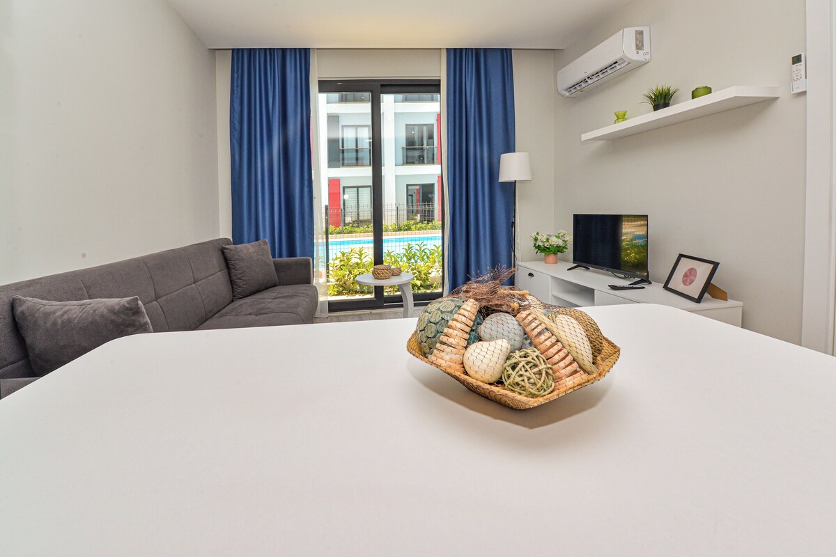 Une chambre d’hôtel moderne avec un canapé gris, une table blanche, un téléviseur et une vue sur une piscine, visible à travers une porte vitrée.
