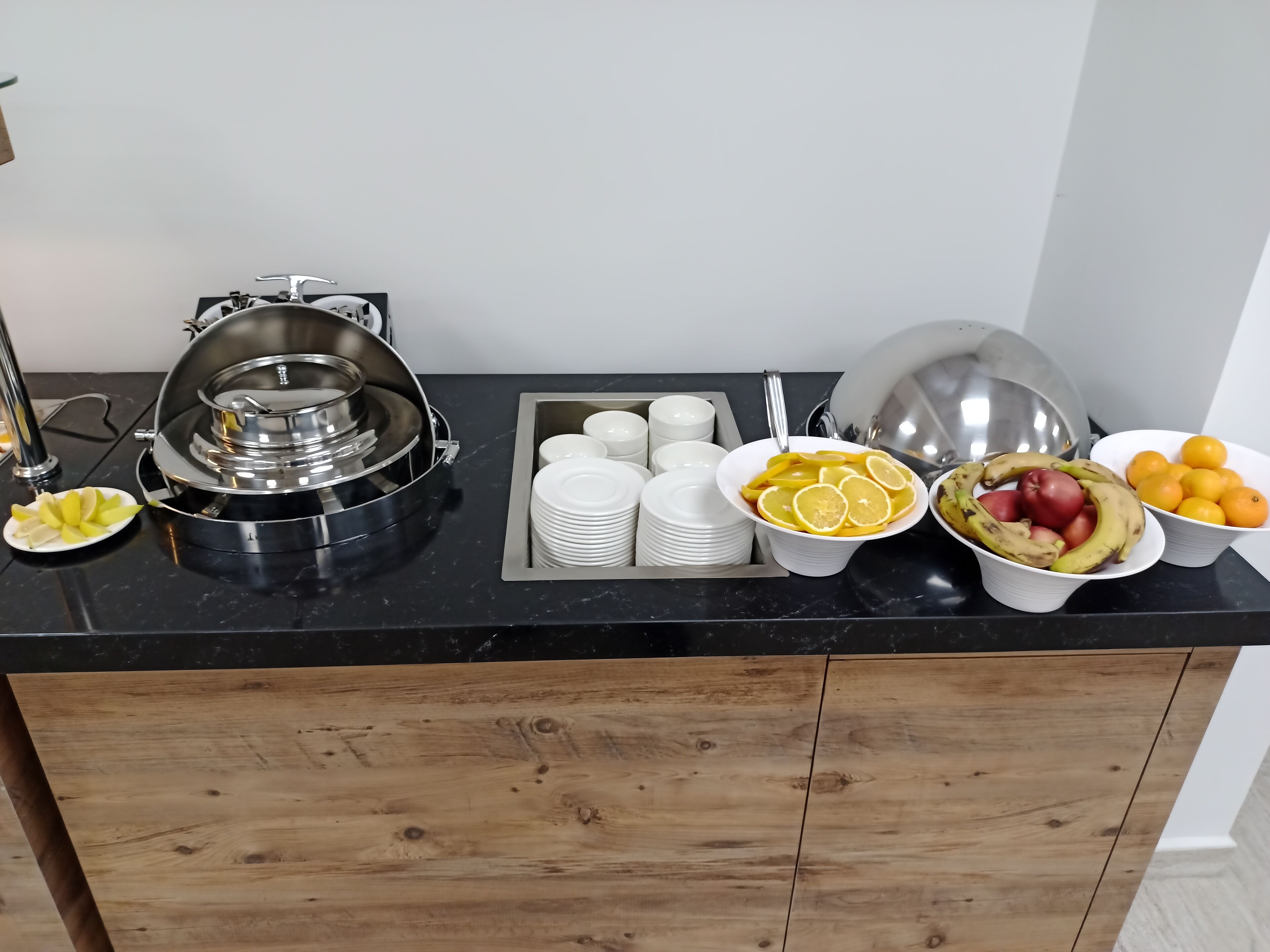 A buffet setup with a self-service hot food station, white plates, bowls of fruit, and a variety of fresh produce.