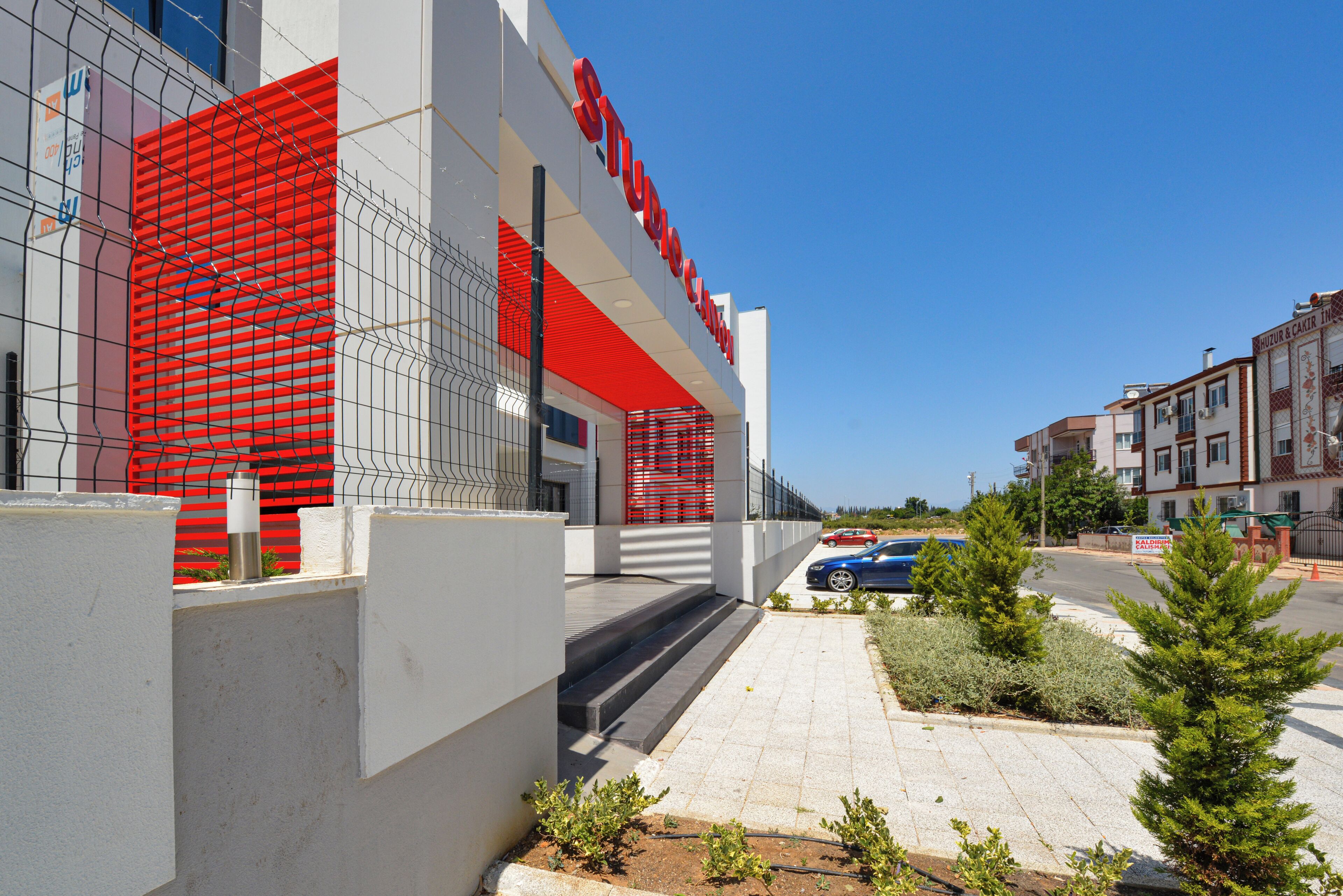 A modern building with a red and white facade, a visible entrance, and a paved area with small plants.